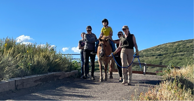 Lucy on a horse being led down an outside path with her therapists.